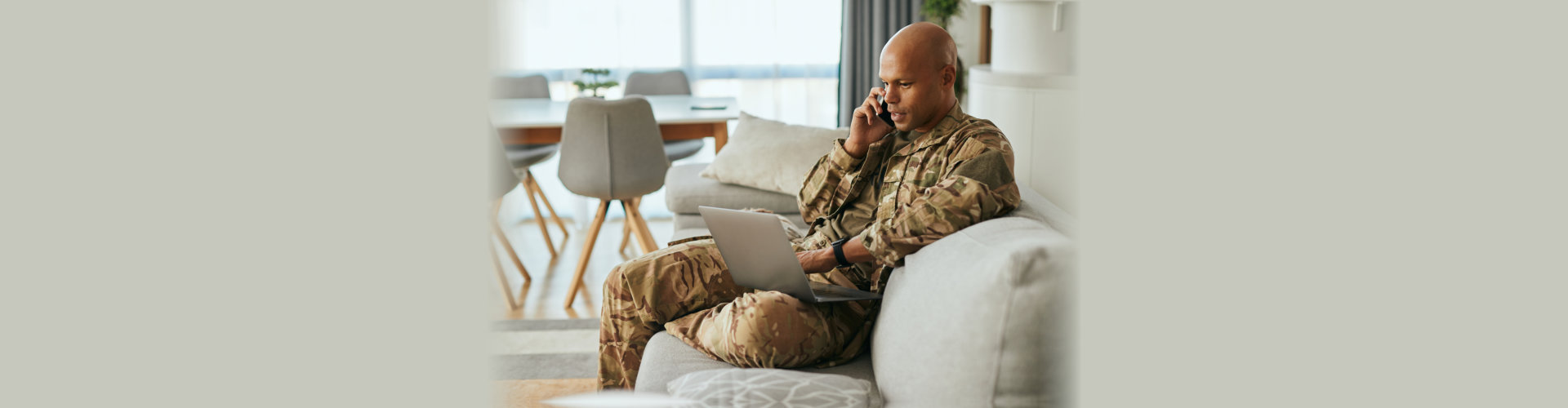 military man uses laptop while talking on cell phone at home